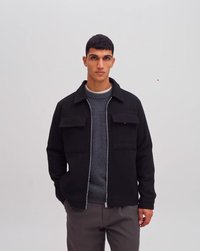 Veste noire zippée avec deux poches avant, confectionnée dans un tissu doux. Portée par-dessus un pull à col ras du cou gris, assortie à un pantalon gris.