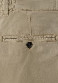 Pantalon kaki beige avec une poche arrière dotée d'une ouverture cousue et d'une fermeture par bouton noir, fabriqué en tissu de coton tissé lisse.