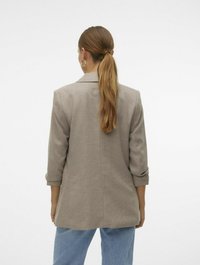 Lys brun blazer med teksturert stoff, skreddersydd passform og rullede ermer, sammen med avslappede blå jeans. Sett fra bak.