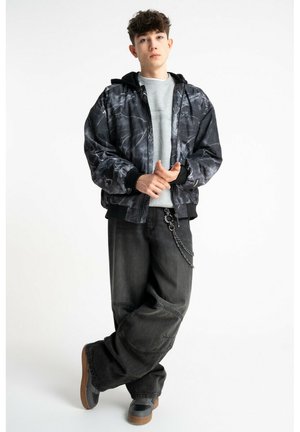 Młody mężczyzna stoi ze złączonymi rękami, ubrany w czarno-szarą kurtkę w wzory, szarą bluzę, luźne czarne spodnie z łańcuchem oraz czarne trampki.