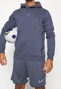 Sweat-shirt à capuche bleu marine avec un accent rouge et un petit logo ; associé à un short assorti. Tenant un ballon de football gris et bleu.