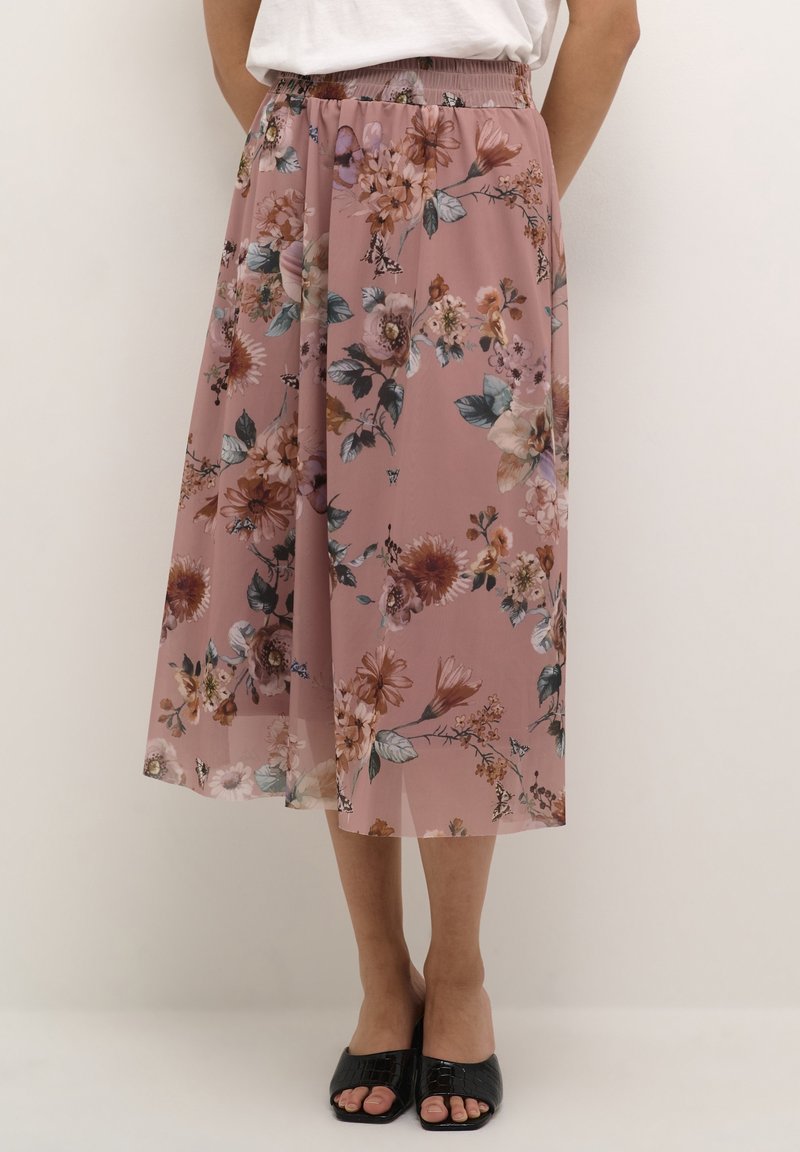 Femme portant une jupe fleurie rose longueur genou avec une taille élastique et des sandales noires ouvertes à l'avant, debout devant un fond uni.