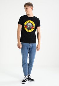 Sort bomulds t-shirt med et cirkulært grafisk design med teksten "GUNS N' ROSES", røde roser og et kranium, parret med lyseblå jeans og sorte sneakers.