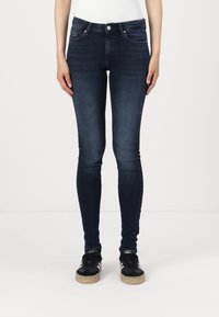 Sötétkék farmer skinny nadrág középmagas derékkal, öt zsebbel és finom koptatott részletekkel az elülső részen. Fekete sportcipővel párosítva.