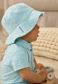 Sombrero de cubo texturizado en azul claro con una camiseta a juego, que presenta un patrón geométrico. Etiqueta "BAKER" en el sombrero. Perfil de un niño visible.