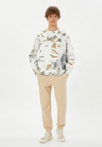 Sudadera de teñido anudado en blanco, beige y gris con mangas largas; combinada con pantalones de chándal beige y zapatillas blancas, con un ajuste relajado.