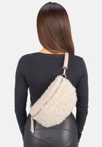 Beiger Shearling-Crossbody-Beutel mit einem Reißverschluss, akzentuiert durch einen glatten Tragegurt und eine strukturierte Oberfläche, diagonal über einem schwarzen Oberteil getragen.