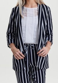 Femme portant un blazer et un pantalon à rayures verticales bleu marine et blanches avec un haut en dentelle blanche, les mains ajustant le devant du blazer.