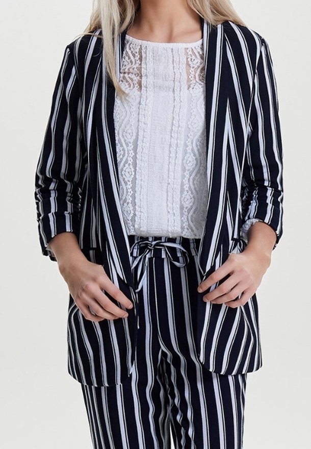 Femme portant un blazer et un pantalon à rayures verticales bleu marine et blanches avec un haut en dentelle blanche, les mains ajustant le devant du blazer.