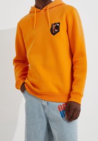 Sudadera con capucha color naranja, con un logo de cabeza de oso en el pecho, que incluye un cordón en la capucha y puños acanalados. Combinada con jeans de color azul claro.