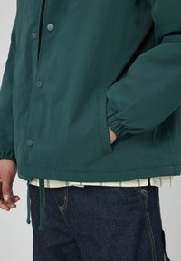 Veste légère verte, au fit décontracté, avec des boutons-pression, des poignets élastiques et un ourlet à cordon de serrage. Portée par-dessus une chemise rayée et un pantalon en denim.