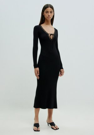 Schwarzes, geripptes Strickkleid mit langen Ärmeln, Spitzenbesatz am Ausschnitt und einer figurbetonten Silhouette, das bis zur Mitte der Waden reicht. Getragen mit schwarzen Absatzsandalen.