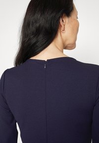 Mörkblå klänning med rund halsringning, som har en synlig dragkedja i ryggen och slät, texturerad tyg.