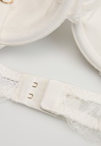 Zbliżenie na biały koronkowy biustonosz z wkładanymi miseczkami i zapięciem na dwa haftki z tyłu na jasnym tle.