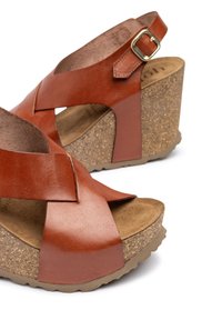 Brown leather wedge sandals featuring a cross-strap design, cork sole, textured footbed, and adjustable ankle strap with a gold buckle.