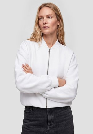 Lyshåret kvinde med bølget hår iført en hvid lynlåstrøje og sorte bukser, stående med armene over kors og kigger til siden.