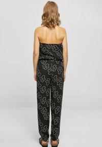 Czarny kombinezon z białym wzorem kwiatowym, bez ramiączek, luźne nogawki, miękka tkanina, a na dole widoczne płaskie sandały.