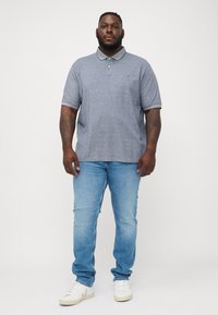 Man med tatueringar som har på sig en grå polo-tröja, ljusblå jeans och vita sneakers, stående mot en helt vit bakgrund.