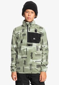 Grüner Fleece-Pullover mit schwarzen Akzenten, halber Reißverschluss und verschiedenen schwarzen geometrischen Mustern, getragen mit einer schwarzen Mütze.