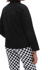 Black, long-sleeve jacket made of cotton, featuring a back yoke and straight cut, paired with black-and-white checkered pants.
