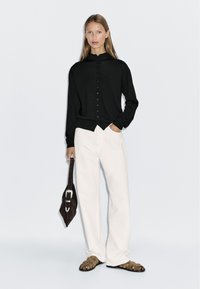 Schwarzes, knopfbares Oberteil mit hohem Kragen, kombiniert mit weißen, locker sitzenden Hosen. Das Model hält eine braune Tasche und steht in Sandalen vor einem schlichten Hintergrund.
