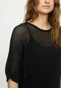 Schwarzes, netzartiges Strickoberteil mit kurzen Ärmeln, weitem Halsausschnitt und lockerer Passform. Die Struktur ist offen und atmungsaktiv.