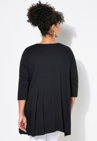 Schwarze Tunika mit dreiviertel Ärmeln, Faltenrücken und lockerer Passform. Aus weichem Stoff gefertigt, hat sie einen runden Halsausschnitt.