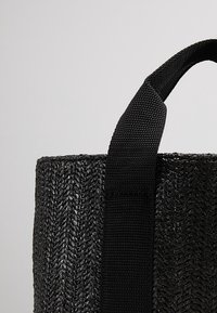 Bolso tote tejido negro con una superficie texturizada similar a la paja, que cuenta con una resistente correa de tela negra para llevarlo.