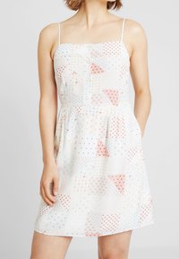 Abito bianco con spalline sottili, caratterizzato da un design a patchwork di motivi rossi e blu. Silhouette svasata con vita arricciata e bottoni.