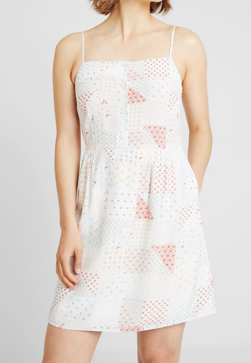 Abito bianco con spalline sottili, caratterizzato da un design a patchwork di motivi rossi e blu. Silhouette svasata con vita arricciata e bottoni.