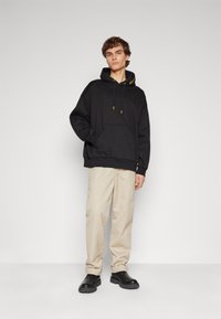 Felpa nera con tasca frontale e lacci gialli, abbinata a pantaloni beige e scarpe nere, su uno sfondo neutro.