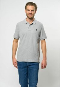 Šedé polo tričko s golierom, krátkymi rukávmi a malým tmavým logom na hrudi. Skombinované s modrými džínsami. Bavlnený materiál, hladká textúra.