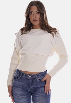 Maglione bianco corto con texture a coste e maniche lunghe, caratterizzato da un colletto a barca e un piccolo dettaglio con logo. Indossato con jeans blu.