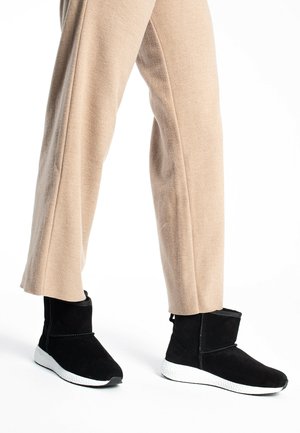 Stivaletti alla caviglia in suede nero con suola bianca, caratterizzati da una texture morbida, punta arrotondata e design semplice, abbinati a pantaloni beige a gamba larga.