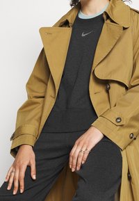 Light brown trench coat with a classic design, button accents, and a smooth texture, layered over a dark sweatshirt with a logo.