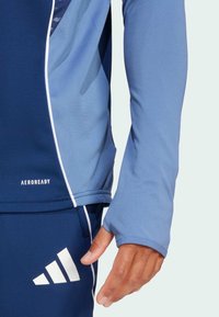 Blauwe atletische langarmtop met een contrasterend lichterblauw paneel, gemaakt van een hoogwaardig functioneel materiaal en met duimgatdetail bij de manchetten.