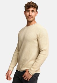 Chemise beige à manches longues avec un col rond. Le tissu présente un motif de tissage subtil et un bord contrastant clair aux poignets.