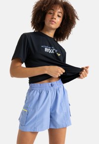 Camisa negra de manga corta con texto de logo en blanco, combinada con pantalones cortos azul claro que tienen una cinturilla elástica y un detalle de bolsillo con cremallera amarilla.