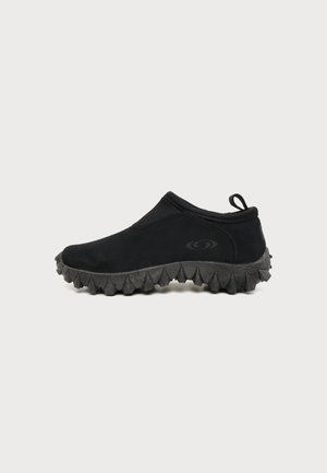 Chaussure noire à enfiler avec une semelle épaisse et dentelée ainsi qu'une languette au talon, montrée de profil sur un fond uni.
