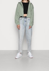 Sweat à capuche court vert avec cordon de serrage, associé à un jean taille haute bleu clair. Des baskets blanches avec des accents roses complètent la tenue.