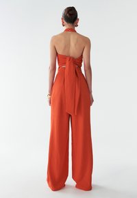 Oranžový dvojdielny outfit so zaviazanou halter blúzkou bez chrbta a širokými nohavicami, prezentujúci štruktúrovanú látku.