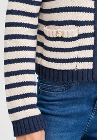 Pletený cardigan s námořnickými a krémovými vodorovnými pruhy, s kapsou na zlatý knoflík, žebrovanými manžetami a lemem, nošený přes modré džíny.