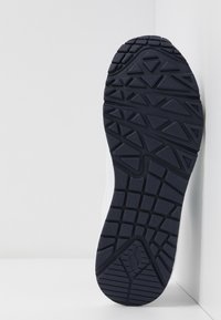 Semelle de basket noire avec motifs géométriques, placée verticalement contre un coin blanc de mur et de sol.