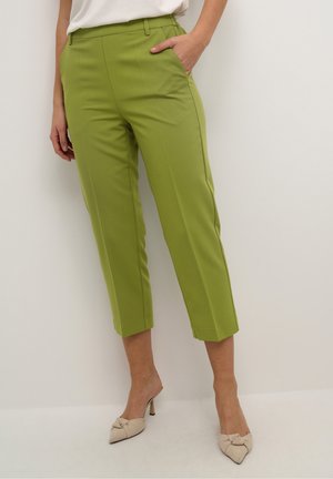 Femme portant un pantalon court vert citron avec des poches latérales et des talons beige pointus avec des nœuds, debout devant un mur clair uni.