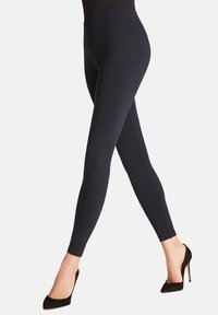 Leggings noirs avec une taille haute, réalisés en tissu lisse et extensible, conçus pour une silhouette ajustée. Portés avec des talons hauts noirs.