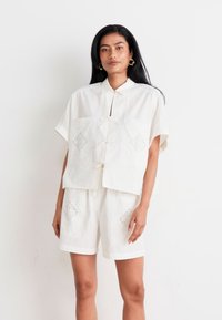 Λευκό βαμβακερό πουκάμισο κοντό μανίκι με διακοσμητικές σχισμές και κλείσιμο με κορδόνι, σε συνδυασμό με ταιριαστά λευκά σορτς που έχουν παρόμοια σχέδια.