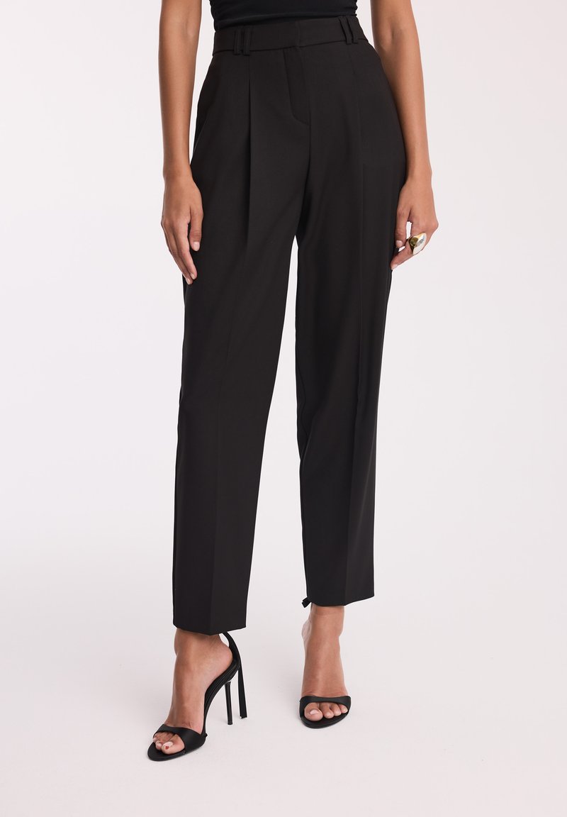 Czarne spodnie z wysokim stanem wykonane z gładkiego materiału, z przodu fałdami, dopasowanym krojem i zwężającą się nogawką. W połączeniu z czarnymi sandałami na obcasie.