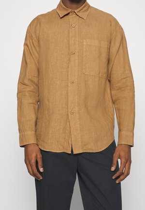 Mężczyzna ubrany w długi rękaw, beżową koszulę na guziki z przednią kieszenią oraz czarne spodnie, stojący na tle jednolitego białego tła.