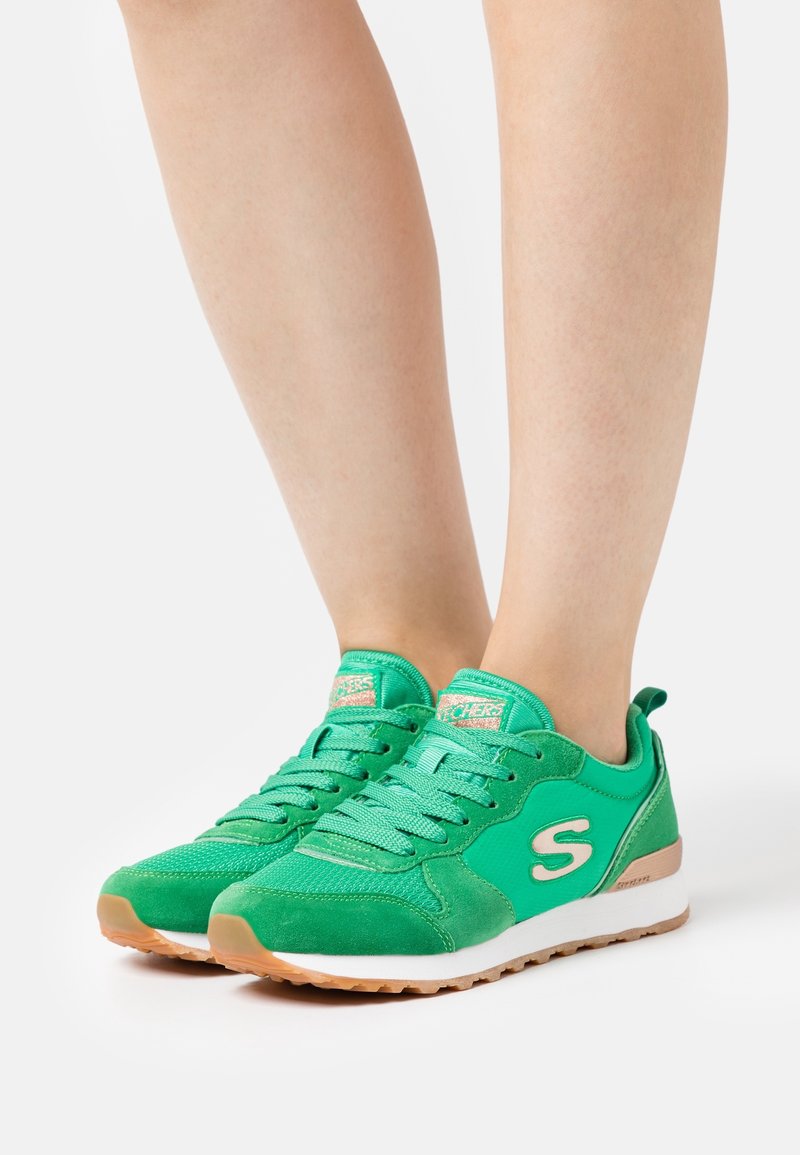 Zapatillas verdes con parte superior de tejido texturizado y detalles en ante, presentando una entresuela blanca y una suela de goma de color goma. Diseño con cordones y detalle de logo.