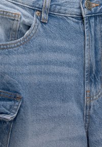 Jeans de mezclilla azul claro con una textura suave, que cuentan con un bolsillo frontal, cierre con botón de metal y detalles de costura en naranja de contraste.